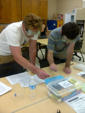 two students in saftey goggles working on a problem togehter as they stand around a table