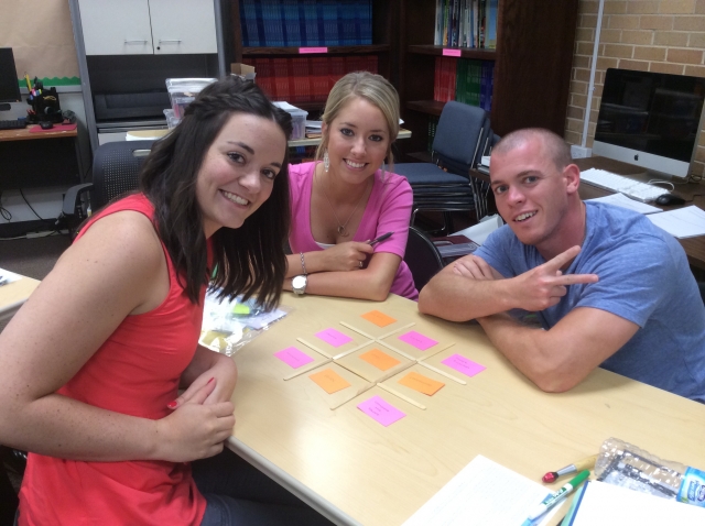 Three students sitting around a table, with sticky notes arranged on the table that mimic a tick tack toe game.  